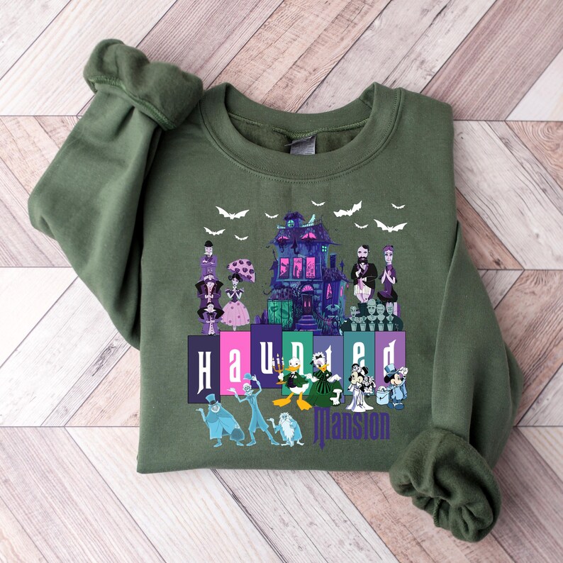 Puede incluir: Una sudadera verde con un estampado gr&aacute;fico de la Mansi&oacute;n Encantada de Disney. El estampado presenta la mansi&oacute;n, fantasmas y el texto "Haunted Mansion".