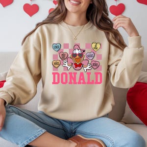 May include: Beige sweatshirt featuring a Valentine's Day graphic of Donald Duck wearing sunglasses and a bow tie. The design includes heart-shaped candies with phrases like "Lover" and "Kisses", and the name "DONALD" in pink block letters.