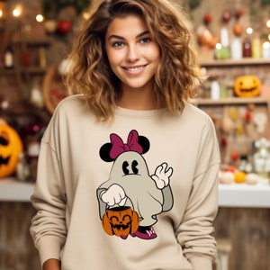 Pode incluir: Camisola bege com um personagem de fantasma de desenho animado com tema de Halloween, com orelhas e la&ccedil;o da Minnie Mouse. O fantasma est&aacute; segurando um balde de ab&oacute;bora. O design usa tons de cinza, preto, rosa e laranja.