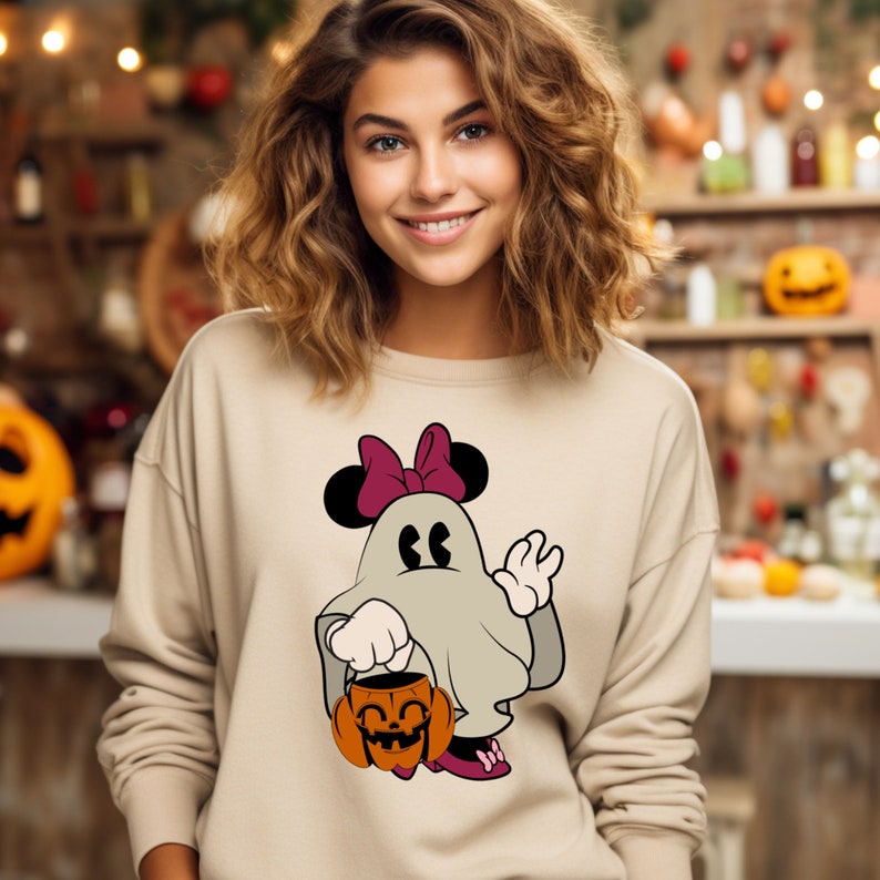 Pode incluir: Camisola bege com um personagem de fantasma de desenho animado com tema de Halloween, com orelhas e la&ccedil;o da Minnie Mouse. O fantasma est&aacute; segurando um balde de ab&oacute;bora. O design usa tons de cinza, preto, rosa e laranja.