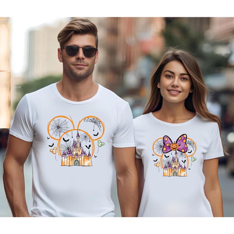 May include: White t-shirts featuring a Halloween-themed design. The design includes a castle, spiderwebs, bats, and a Mickey Mouse head outline. One shirt has a bow with a pumpkin pattern.