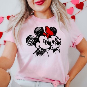 May include: A light pink t-shirt featuring a black and white sketch of Mickey and Minnie Mouse. Minnie Mouse is wearing a red bow. The t-shirt has short sleeves and a crew neck.