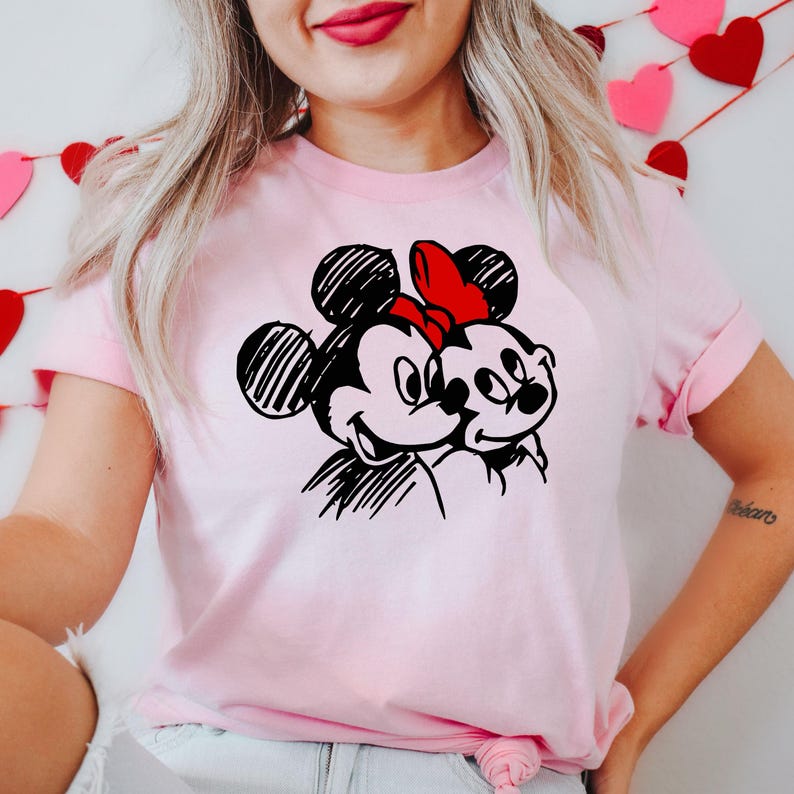 May include: A light pink t-shirt featuring a black and white sketch of Mickey and Minnie Mouse. Minnie Mouse is wearing a red bow. The t-shirt has short sleeves and a crew neck.