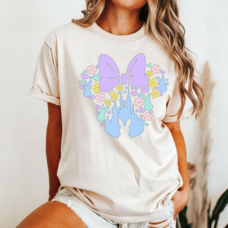 May include: Beige t-shirt featuring a pastel Easter-themed graphic. The design includes a purple bow, a blue castle, flowers, and bunny silhouettes. The shirt has short sleeves and a relaxed fit.