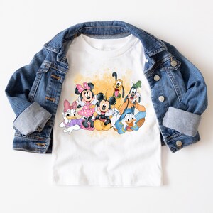 Puede incluir: Camiseta blanca con una ilustraci&oacute;n colorida de Mickey Mouse, Minnie Mouse, Donald Duck, Goofy y Pluto. Los personajes sonr&iacute;en y parecen estar divirti&eacute;ndose juntos.