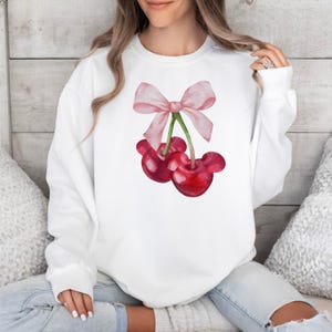 Puede incluir: Sudadera blanca con un lazo rosa y dos cerezas rojas en la parte delantera.