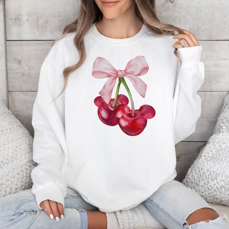 Puede incluir: Sudadera blanca con un lazo rosa y dos cerezas rojas en la parte delantera.
