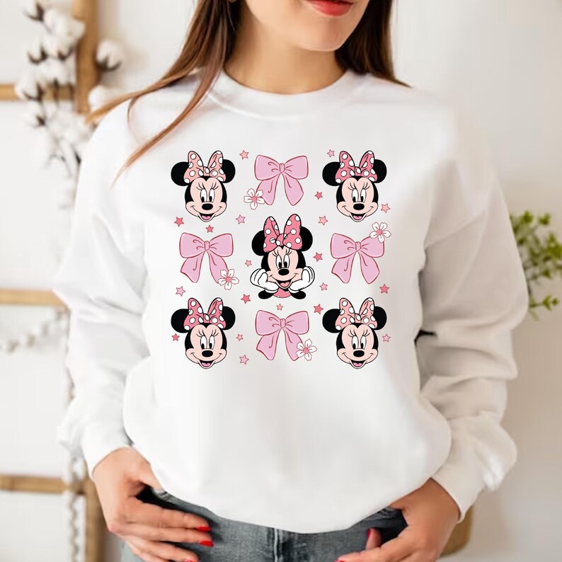 May include: White sweatshirt with a pink and red Minnie Mouse print. The print features Minnie Mouse's face with a pink bow and polka dots. The print is repeated multiple times on the sweatshirt.
