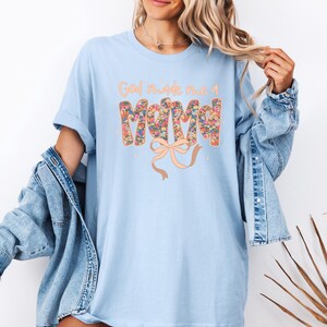 Pu&ograve; includere: T-shirt azzurra con la scritta "God made me a Mama" in un design floreale. La parola "Mama" &egrave; riempita con un motivo floreale colorato e un fiocco color pesca. La maglietta &egrave; indossata con una giacca di jeans.