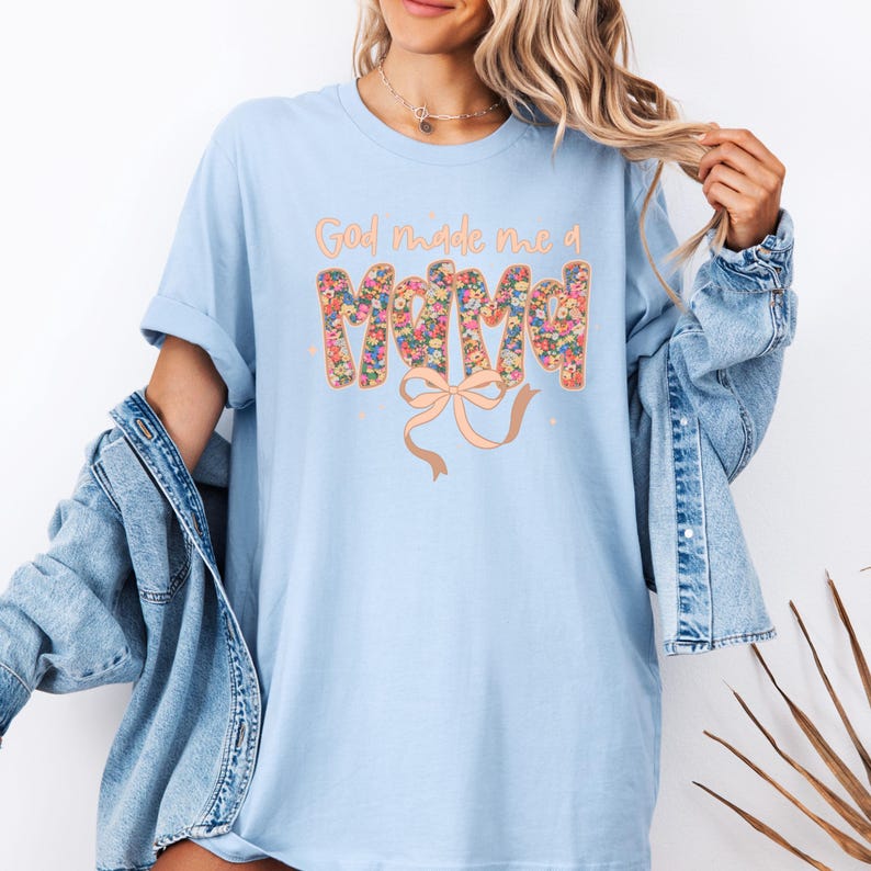 Pu&ograve; includere: T-shirt azzurra con la scritta "God made me a Mama" in un design floreale. La parola "Mama" &egrave; riempita con un motivo floreale colorato e un fiocco color pesca. La maglietta &egrave; indossata con una giacca di jeans.