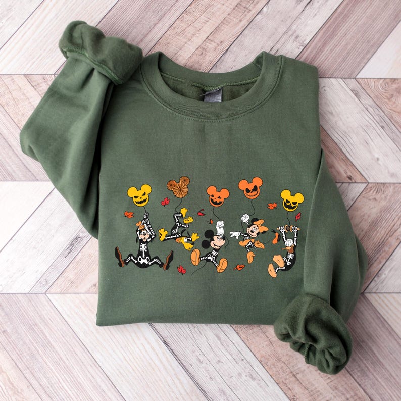 May include: An olive green sweatshirt with a Halloween design. The graphic shows cartoon characters as skeletons, holding Mickey Mouse-shaped balloons in orange and yellow. The design includes autumn leaves.