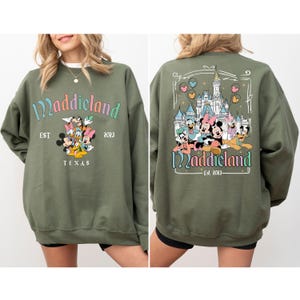 May include: A green sweatshirt with a graphic of Mickey Mouse and friends in front of a castle. The text "Maddieland Est. 2023" is printed on the sweatshirt.