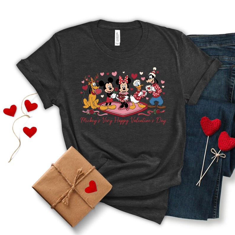 May include: Dark gray t-shirt featuring Disney characters Pluto, Mickey, Minnie, Donald, and Goofy on a pink heart-shaped rug. The text "Mickey's Very Happy Valentine's Day" is printed below the characters. The shirt is paired with blue jeans and Valentine's Day decorations.