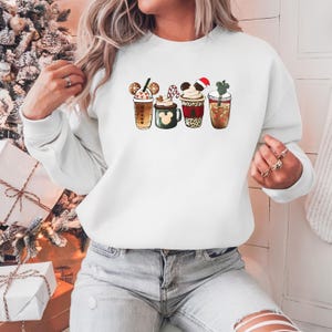 May include: White sweatshirt featuring four festive drink designs. The drinks include a gingerbread cookie, a Mickey Mouse-shaped mug, a Santa hat, and a cactus. The sweatshirt is a comfortable, casual style, perfect for the holiday season.