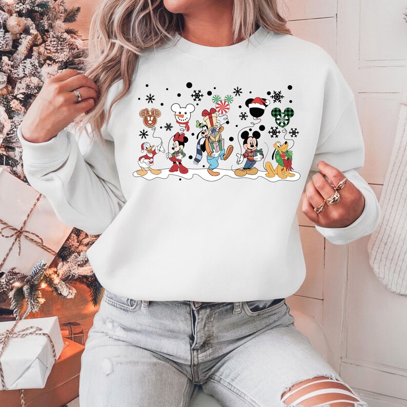 May include: White Christmas-themed sweatshirt featuring a graphic print of Mickey Mouse and his friends, including Donald Duck, Goofy, Pluto, and a snowman. The characters are wearing festive attire and holding gifts.