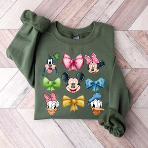 May include: A green sweatshirt with a variety of Disney characters and bows printed on the front. The characters include Mickey Mouse, Minnie Mouse, Donald Duck, Goofy, and Daisy Duck.