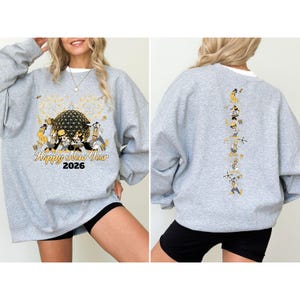 May include: A light gray sweatshirt with a graphic design featuring a large sphere, fireworks, and cartoon characters. The text "Happy New Year 2026" is printed below the graphic. The back of the sweatshirt has a vertical line of cartoon characters.