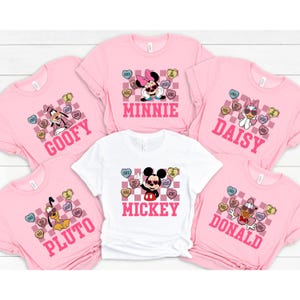 May include: Pink and white t-shirts featuring Disney characters Mickey, Minnie, Goofy, Pluto, and Daisy. Each shirt has a character name and a heart-shaped candy graphic with phrases like "Love" and "Hugs".