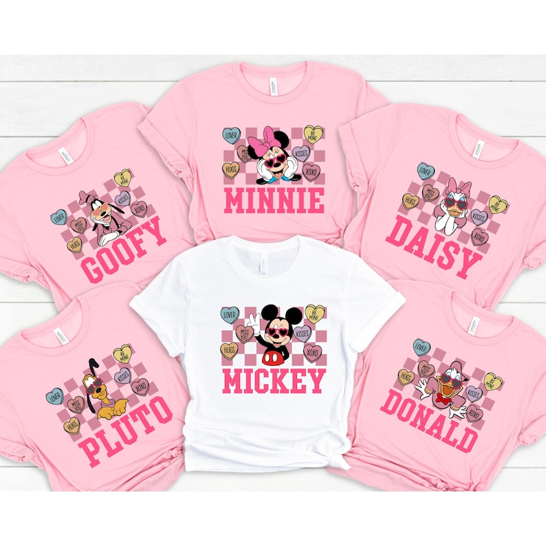 May include: Pink and white t-shirts featuring Disney characters Mickey, Minnie, Goofy, Pluto, and Daisy. Each shirt has a character name and a heart-shaped candy graphic with phrases like "Love" and "Hugs".