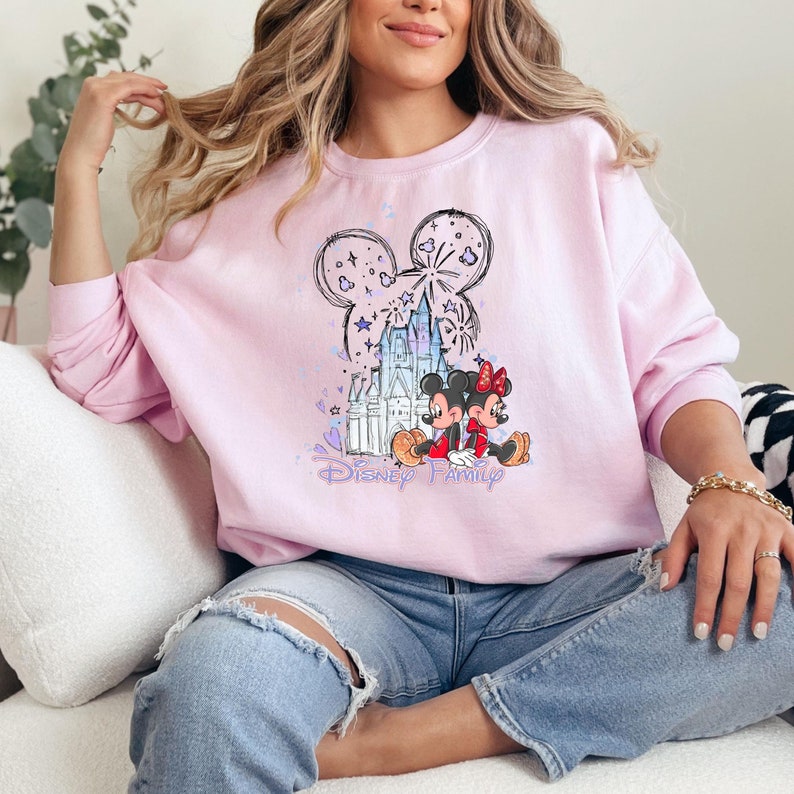 May include: A light pink sweatshirt with a Disney Family design. The graphic features Mickey and Minnie Mouse, a castle, fireworks, and the words "Disney Family" in purple script. The sweatshirt has long sleeves.