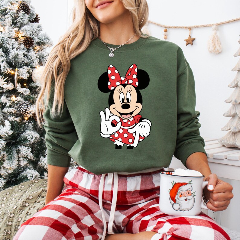 May include: Green sweatshirt with a Minnie Mouse graphic in a red polka dot dress and bow. The cartoon character is making an "okay" hand gesture. The person is wearing red and white plaid pyjama bottoms and holding a mug with a Santa Claus design.