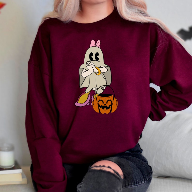 Puede incluir: Una sudadera color burdeos con un fantasma blanco que lleva un lazo rosa y sostiene un cubo de calabaza. El fantasma tiene una expresi&oacute;n de sorpresa y se est&aacute; tapando la cara con las manos.