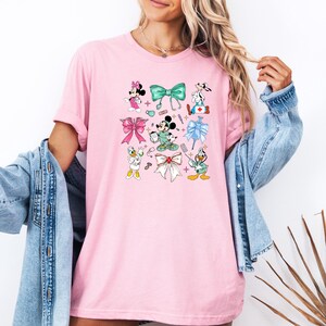 Pu&ograve; includere: T-shirt rosa chiaro con personaggi dei cartoni animati e fiocchi. Il design include Minnie Mouse, Mickey Mouse, Paperino e Pippo, insieme a elementi a tema medico e vari disegni di fiocchi in diversi colori. La maglietta &egrave; di colore rosa chiaro.