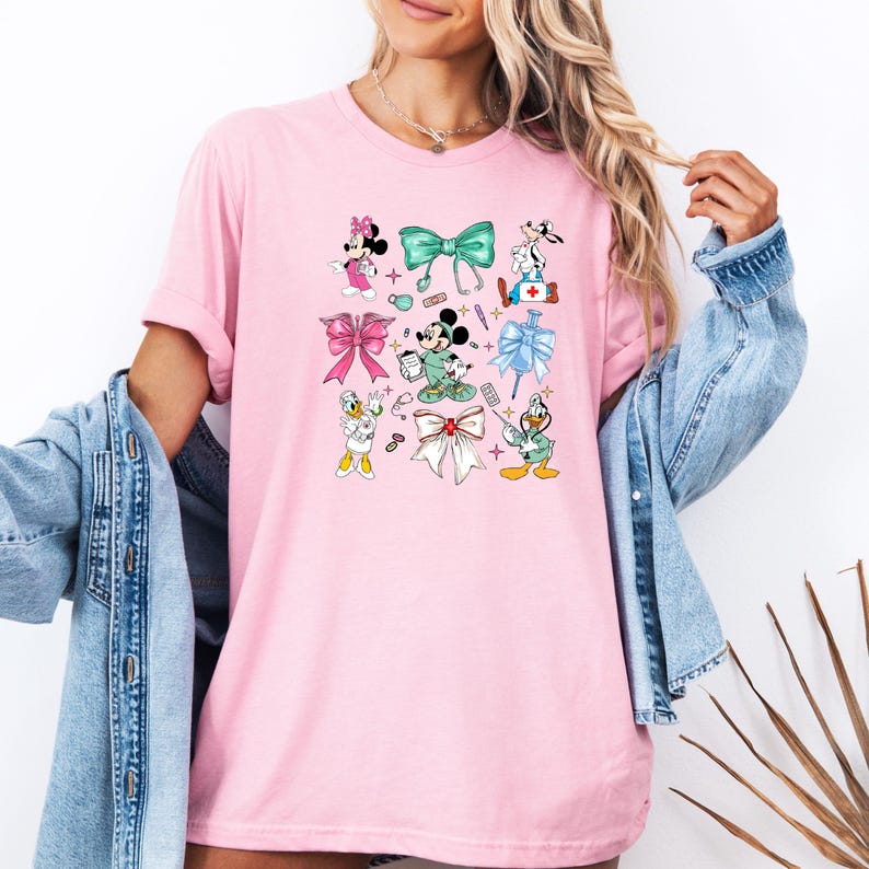 Pu&ograve; includere: T-shirt rosa chiaro con personaggi dei cartoni animati e fiocchi. Il design include Minnie Mouse, Mickey Mouse, Paperino e Pippo, insieme a elementi a tema medico e vari disegni di fiocchi in diversi colori. La maglietta &egrave; di colore rosa chiaro.