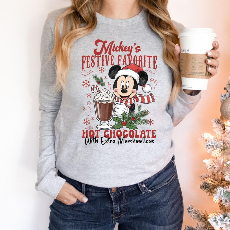 May include: A gray long-sleeve shirt featuring Mickey Mouse in a Santa hat and scarf, holding a cup of hot chocolate with marshmallows. The text reads "Mickey's Festive Favorite Hot Chocolate With Extra Marshmallows."