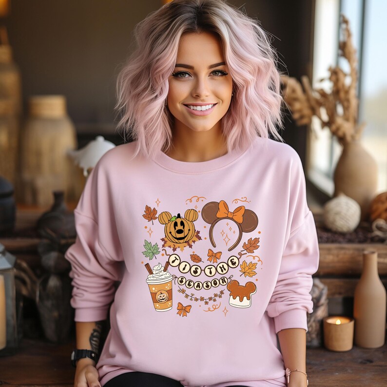 May include: A light pink sweatshirt featuring a fall-themed graphic. The design includes a pumpkin with Mickey Mouse ears, a headband with a bow, fall leaves, a coffee cup, and the text "'Tis the Season".