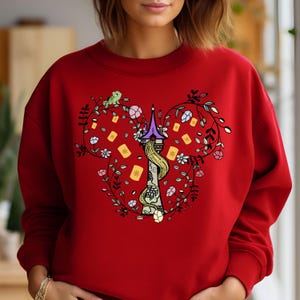 May include: Red sweatshirt with a floral design featuring a tower, a chameleon, and a braid of hair. The design is in the shape of Mickey Mouse ears.