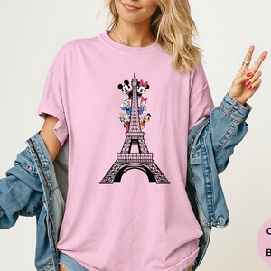 May include: A light pink t-shirt featuring a black graphic of the Eiffel Tower with Mickey Mouse, Minnie Mouse, Donald Duck, Daisy Duck, and Pluto. The text "Comfort Colors Blossom" is in a pink circle.