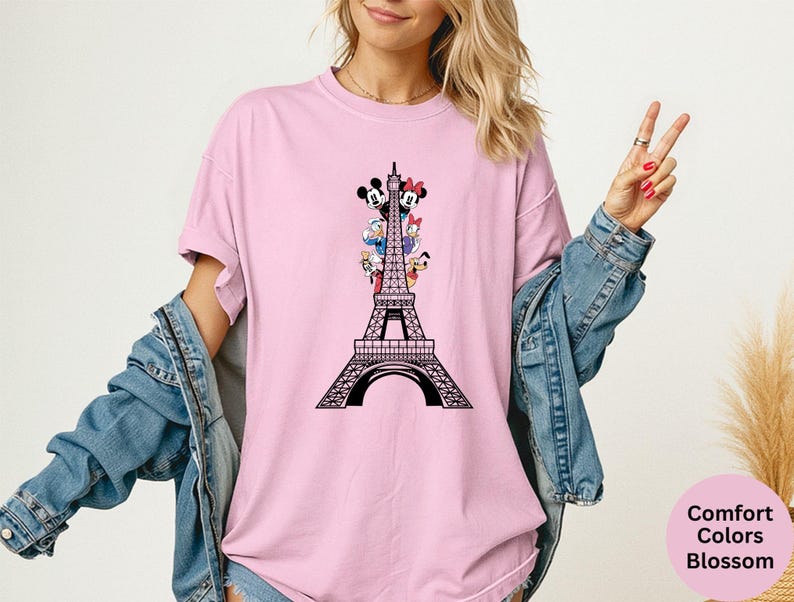 May include: A light pink t-shirt featuring a black graphic of the Eiffel Tower with Mickey Mouse, Minnie Mouse, Donald Duck, Daisy Duck, and Pluto. The text "Comfort Colors Blossom" is in a pink circle.