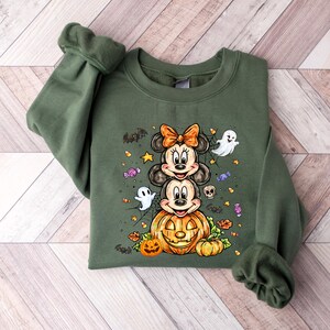 May include: Green sweatshirt with a Halloween design. It features Mickey and Minnie Mouse stacked on a carved pumpkin, with ghosts, bats, and sweets. The sleeves are cuffed.