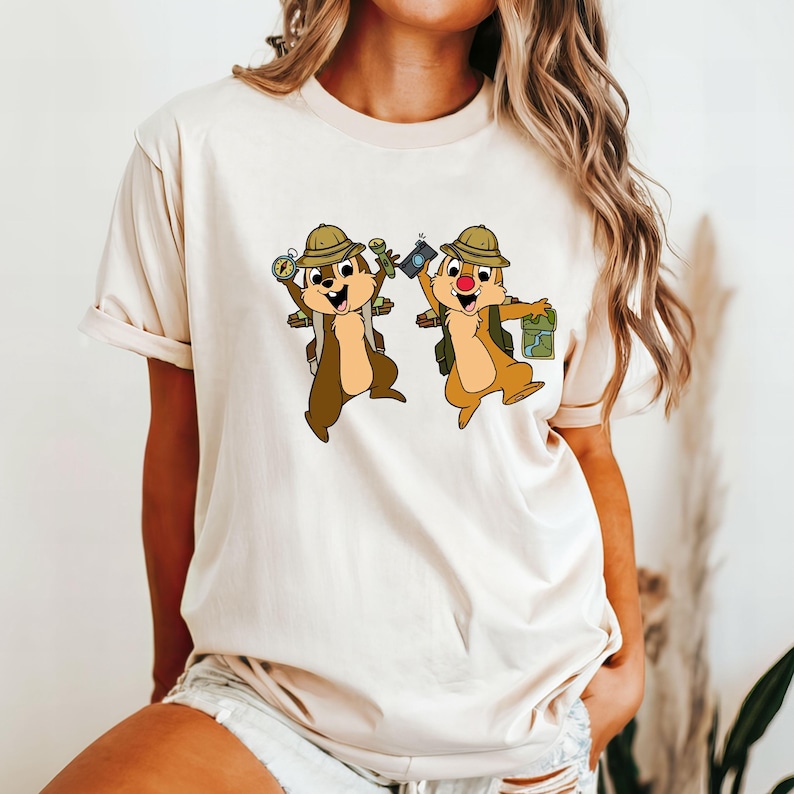 May include: A beige t-shirt featuring a graphic of two cartoon chipmunks wearing explorer hats and carrying camping equipment. The chipmunks are brown with white bellies and are smiling.