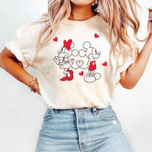 Op de afbeelding: Een wit T-shirt met een afbeelding van Mickey Mouse en Minnie Mouse die hand in hand staan met rode hartjes. De afbeelding is in zwart-wit met rode accenten.