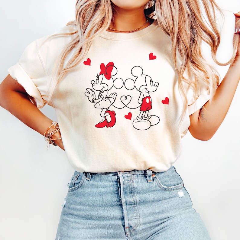 Op de afbeelding: Een wit T-shirt met een afbeelding van Mickey Mouse en Minnie Mouse die hand in hand staan met rode hartjes. De afbeelding is in zwart-wit met rode accenten.