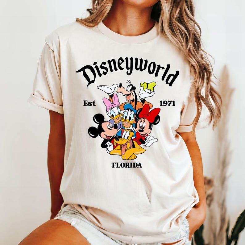 Pu&ograve; includere: Una maglietta beige con una grafica di Topolino, Minnie Mouse, Paperino, Pippo e Pluto. La grafica &egrave; circondata dal testo "Disneyworld Est 1971 Florida".