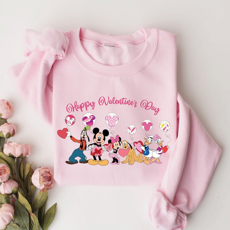 May include: Pink sweatshirt with a graphic of Mickey Mouse and friends holding heart-shaped balloons. The text "Happy Valentine's Day" is printed above the graphic.