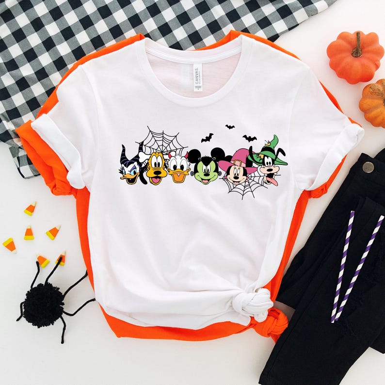 Pu&ograve; includere: Maglietta bianca con una grafica di Topolino, Minnie Mouse, Paperino, Pippo e un personaggio di strega davanti a una ragnatela. I personaggi indossano tutti costumi di Halloween.