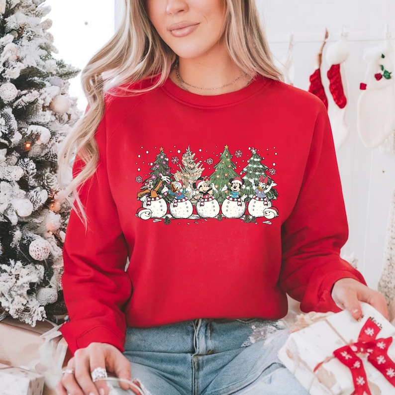 Op de afbeelding: Rode kersttrui met een groep Disney-figuren die verkleed zijn als sneeuwpoppen voor versierde kerstboom. De figuren zijn Mickey Mouse, Minnie Mouse, Donald Duck, Goofy en Pluto.