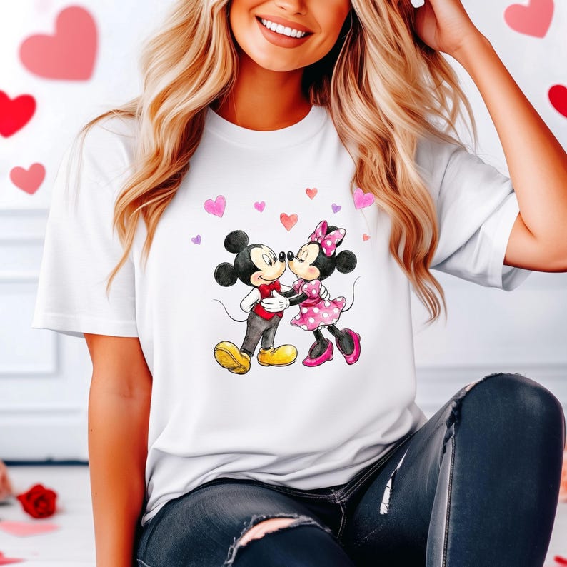 May include: White t-shirt with a watercolour illustration of Mickey and Minnie Mouse kissing, surrounded by pink and purple hearts. Mickey wears a red waistcoat and black trousers, while Minnie has a pink polka-dot dress. A romantic design for Valentine's Day.