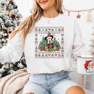 May include: White Christmas-themed sweatshirt featuring Mickey Mouse wearing a Santa hat, surrounded by presents and Christmas trees. The design includes a festive pattern of trees, snowflakes, and hearts. A mug with a Santa Claus design is also visible.