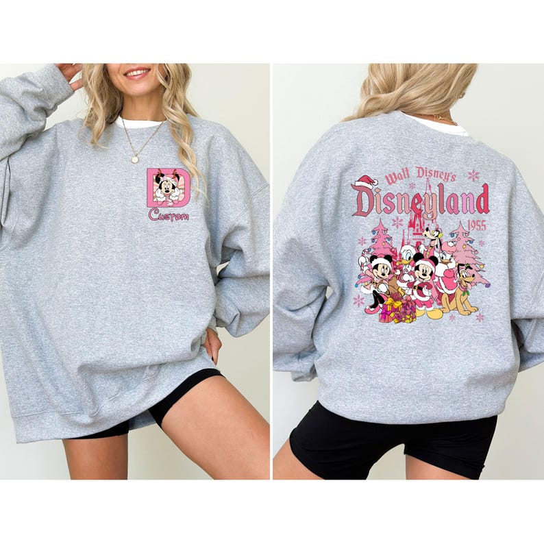 May include: A light gray sweatshirt with a white collar. The front features a pink square with a Minnie Mouse graphic and the word "Custom". The back displays "Walt Disney's Disneyland" with a group of Disney characters and a Christmas tree design.
