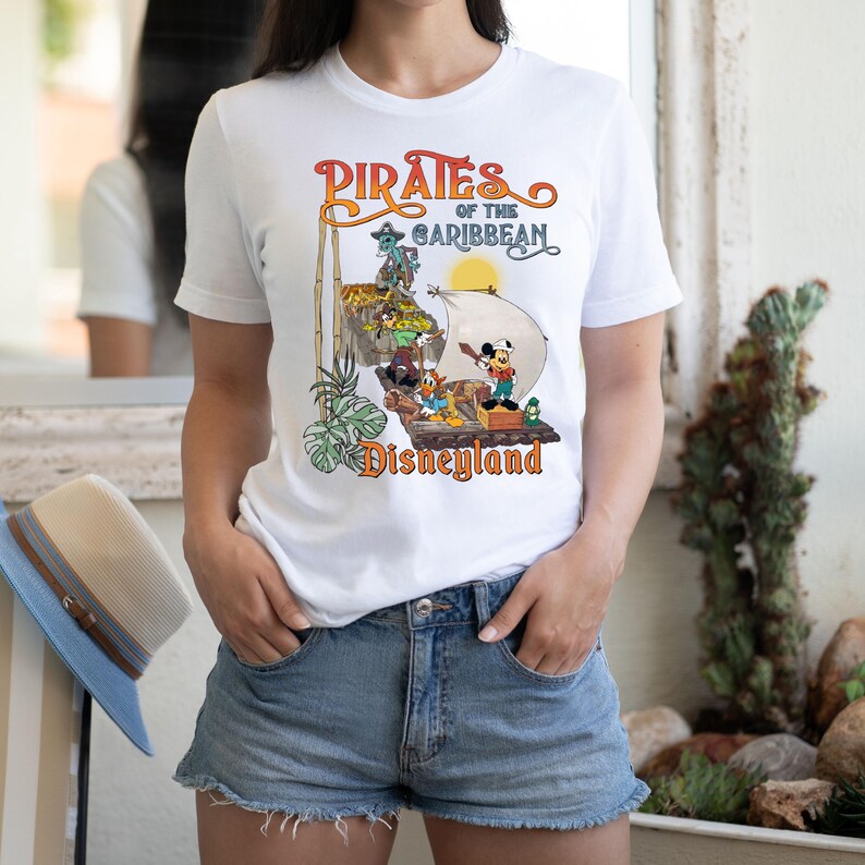 May include: White t-shirt with a vintage-style graphic of Mickey Mouse and other Disney characters on a pirate ship. The text "Pirates of the Caribbean Disneyland" is printed on the shirt.