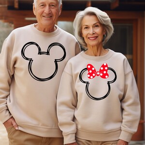 May include: A couple wearing matching tan sweatshirts with a black outline of Mickey Mouse's head. The woman's sweatshirt has a red bow with white polka dots on top of Mickey's head.