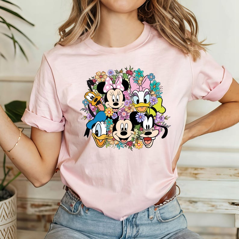 May include: Pink t-shirt with a colorful graphic of Mickey Mouse, Minnie Mouse, Donald Duck, Goofy, and Pluto surrounded by flowers.