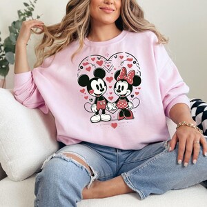 May include: A pink sweatshirt with a graphic of Mickey Mouse and Minnie Mouse holding hands inside a heart. The heart is surrounded by small red hearts and black lines.