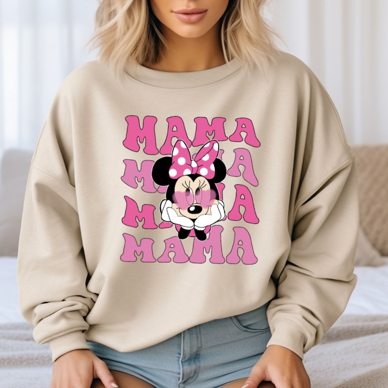 May include: A beige sweatshirt with a pink graphic of Minnie Mouse wearing a pink bow and heart-shaped sunglasses. The text "MAMA" is repeated three times in pink.