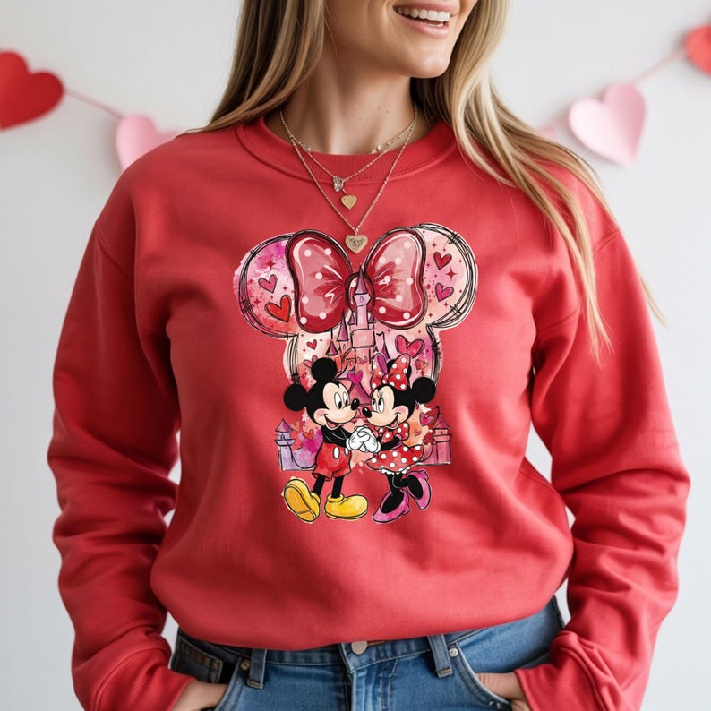 May include: Red sweatshirt featuring a graphic of Mickey and Minnie Mouse embracing in front of a castle. The design includes a large bow with hearts and a polka dot pattern. The sweatshirt is a solid red color.